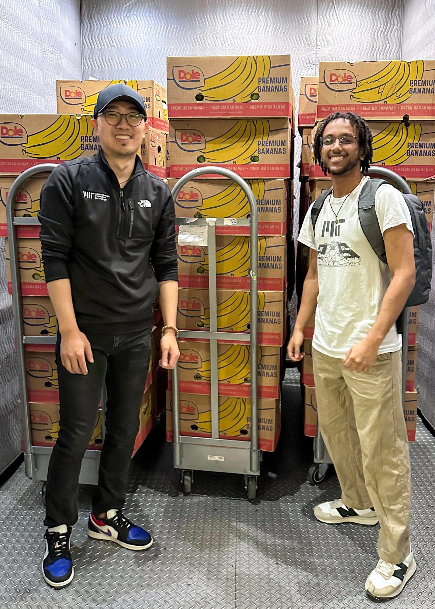 Two people standing in an elevator with carts loaded with boxes of bananas