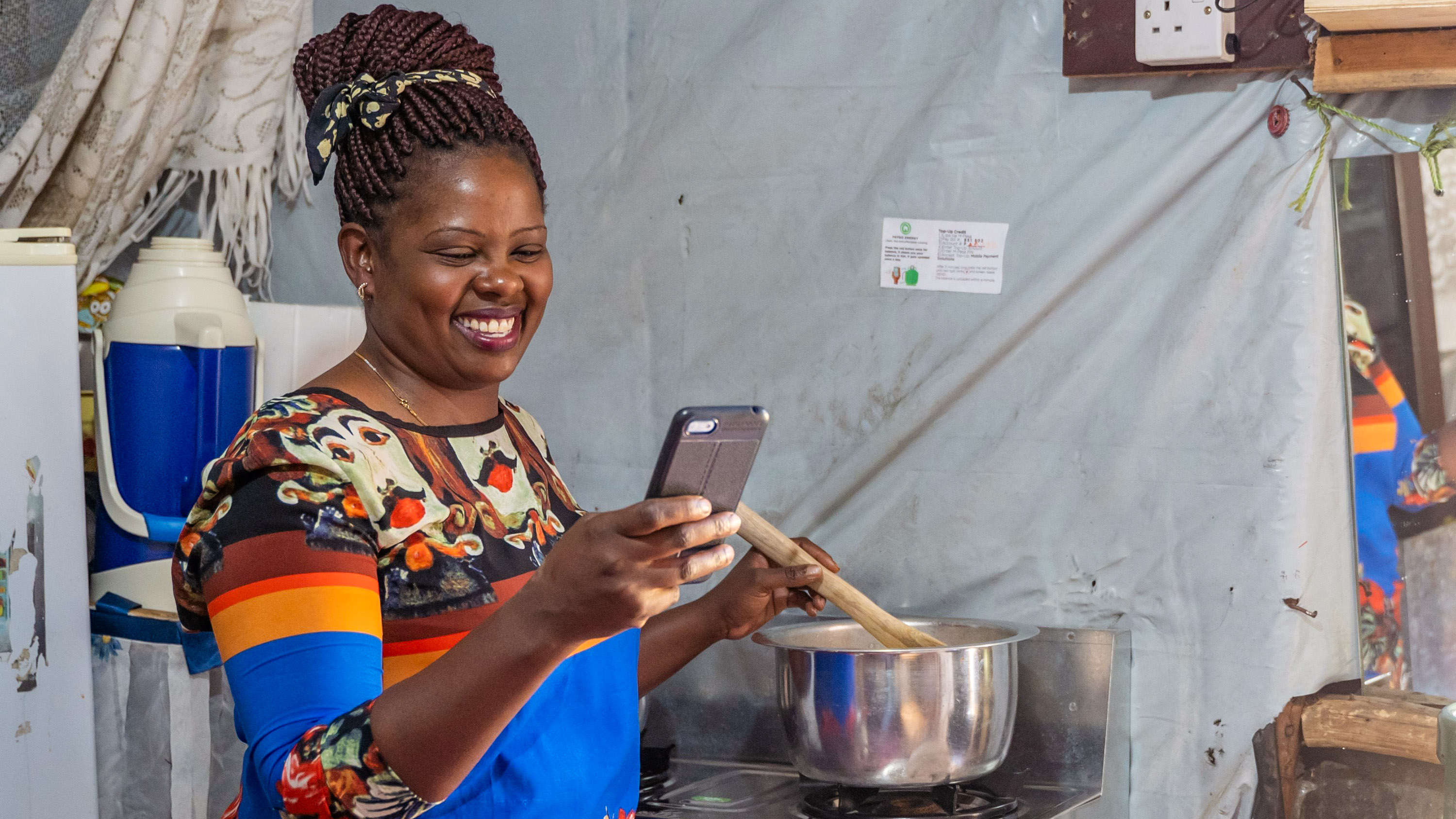 a woman smiling at her phone while stirring a pot on a cookstove burner